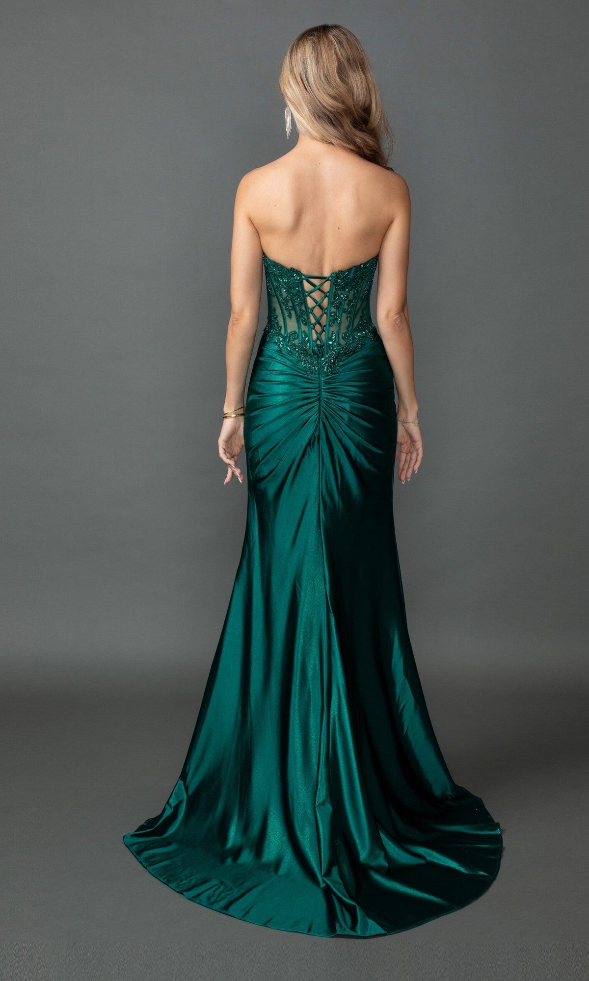 A woman wearing the Long Prom Dress: Dancing Queen 4560—a strapless dark green gown with a lace-up back and subtle ruching—stands with her back to the camera against a plain gray background.